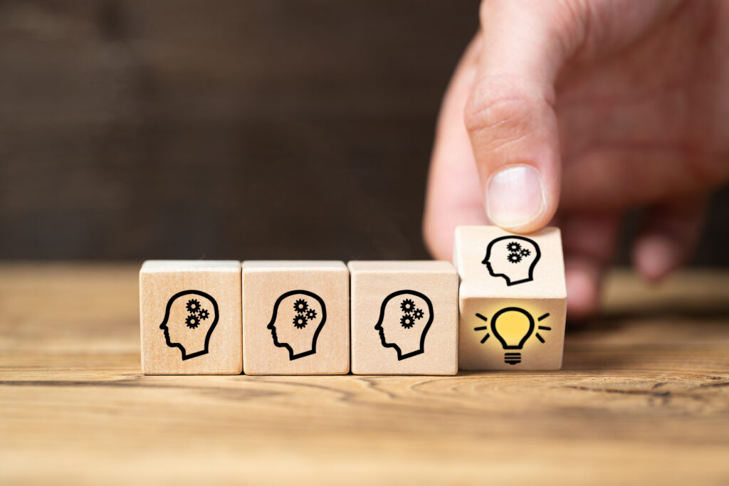 Three wooden blocks with images of a head with cogs inside. Fourth wooden block with same image on it being moved by a hand, revealing a second image of a lit lightbulb