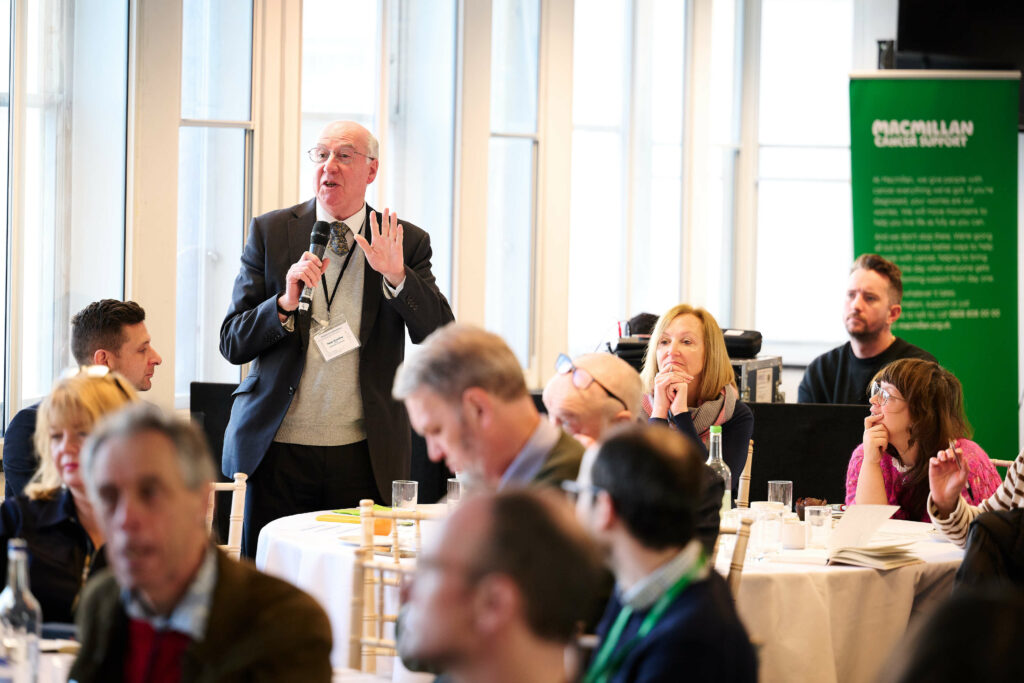 Peter standing at table with microphone in hand speaking to rest of workshop attendees.