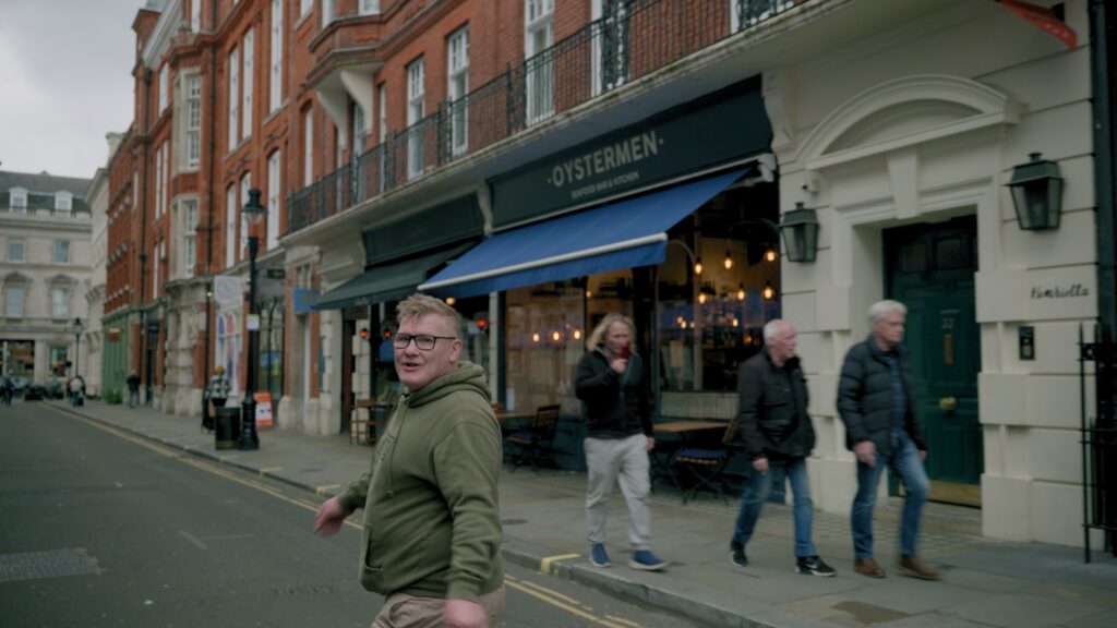 Searching For Answers Still - a man wearing glasses and a hooded top walking down a street looking back at the camera
