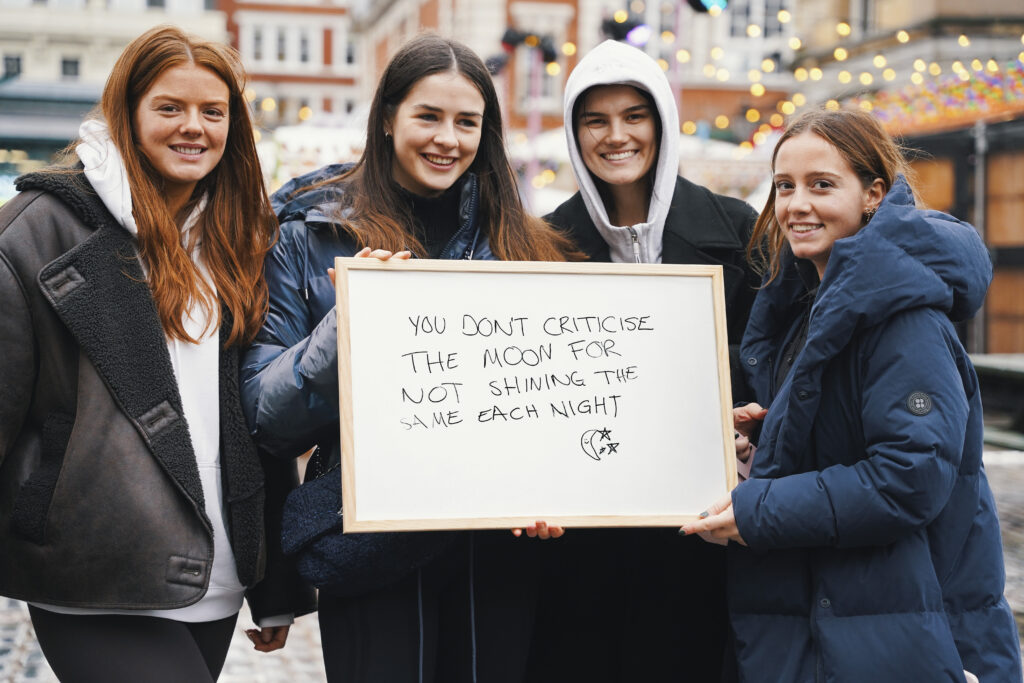 A group of four young women holding a whiteboard displaying the message "you don't criticise the moon for not shining the same each night"