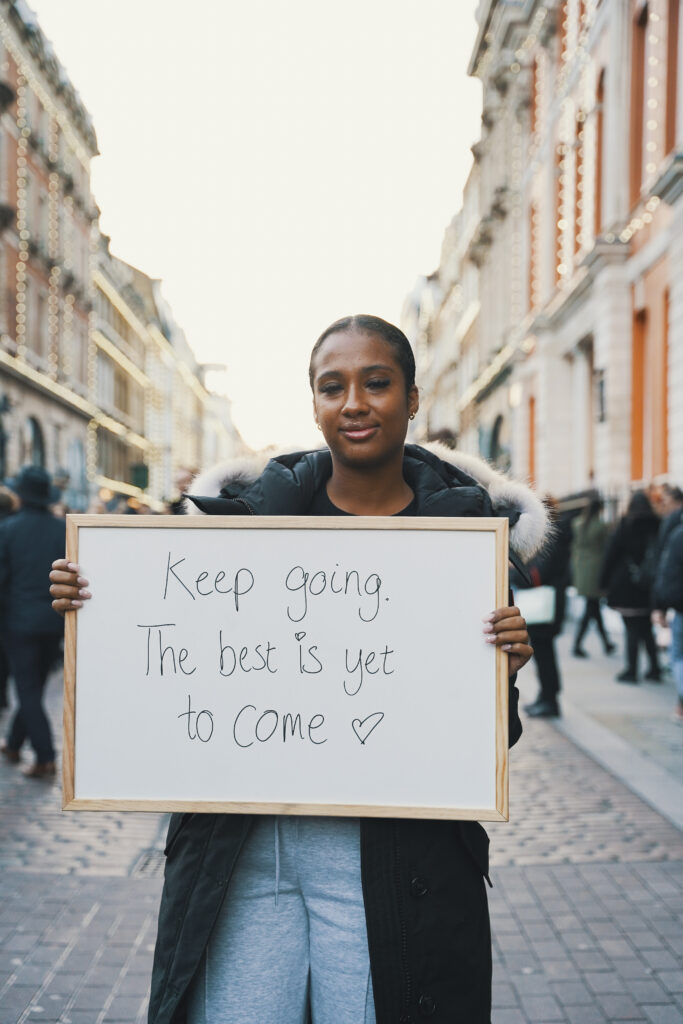 A young woman holding a whiteboard displaying the message "keep going, the best is yet to come"