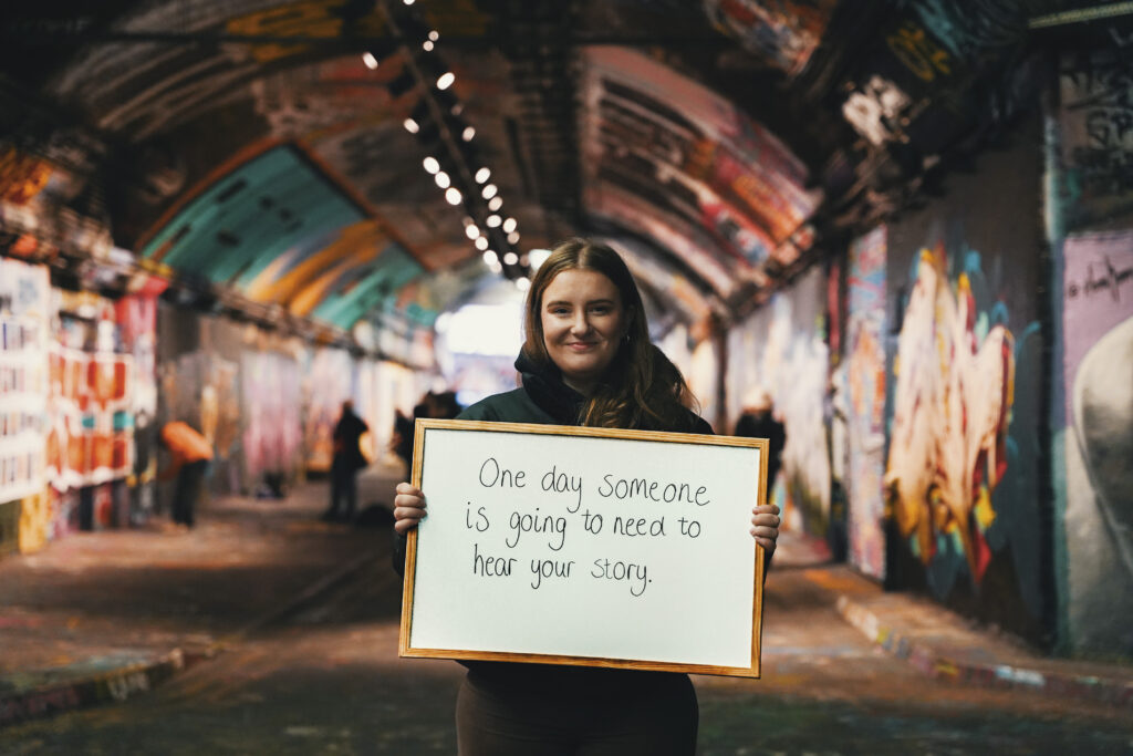 A young woman holding a whiteboard displaying the message "one day someone is going to need to hear your story"
