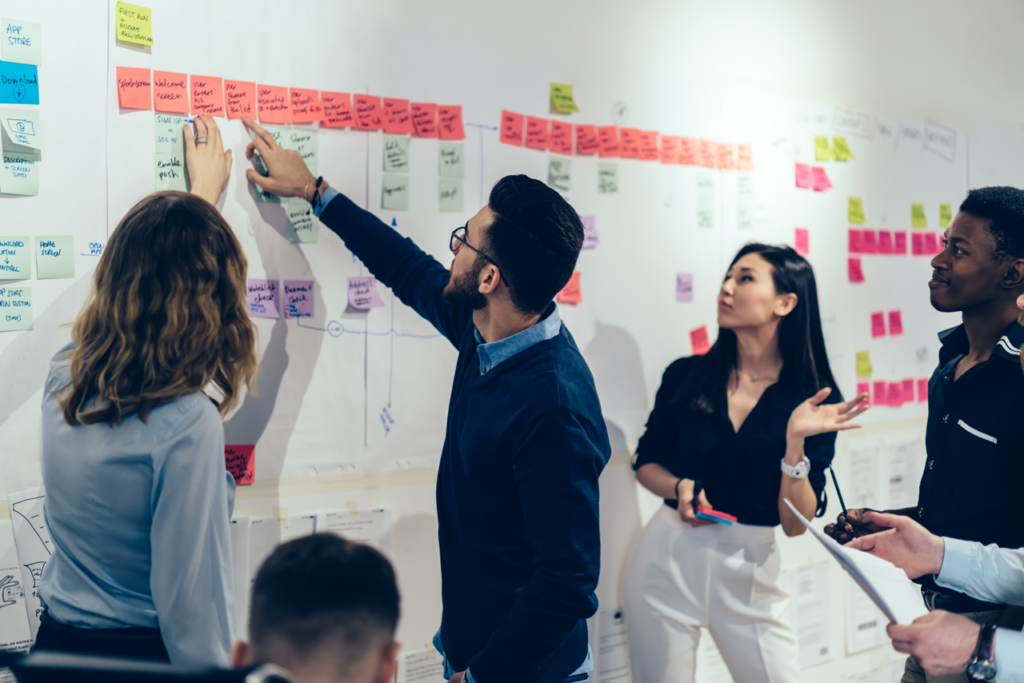 A male and female presenting person pointing to a post it flow diagram on a wall