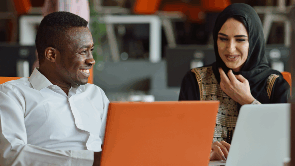 Two people, one male presenting from the global majority and one female presenting with a head scarf smiling whilst looking at laptops