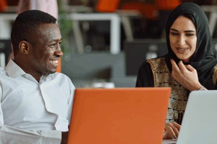 Two people, one male presenting from the global majority and one female presenting with a head scarf smiling whilst looking at laptops