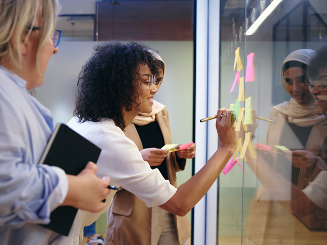 Three colleagues collaborating on a process mapping exercise, writing on a glass wall covered in colourful sticky notes.