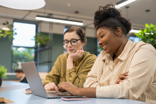 Two colleagues collaborating at a laptop in a modern office, one pointing at the screen as the other smiles.