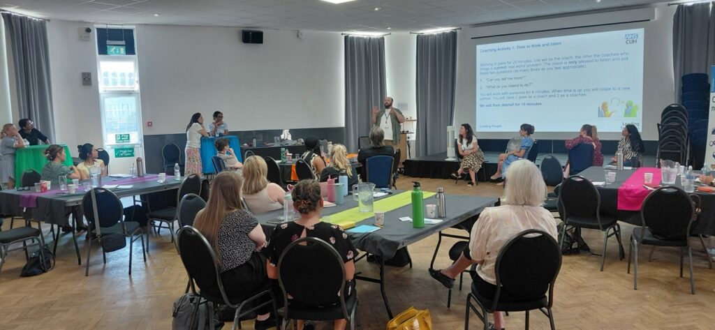 A group of around 25 people take part in a training workshop in a bright conference room. Participants are seated around tables arranged in a semi-circle while a facilitator stands at the front, speaking and gesturing beside a projected presentation slide. Several attendees are seated in a discussion circle near the screen, with notebooks, reusable water bottles and refreshments on the tables, creating a collaborative learning environment.