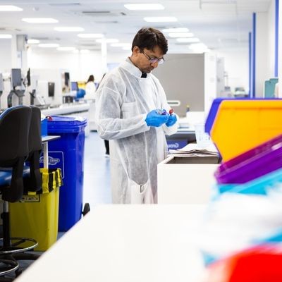 A laboratory worker wearing a white disposable gown, blue gloves and safety glasses stands at a bench, carefully examining a small sample or vial. The modern laboratory is bright and spacious, with equipment and workstations stretching into the background. Colour-coded clinical waste bins are visible nearby, while blurred laboratory containers in the foreground frame the scene, highlighting the focus on careful scientific work.