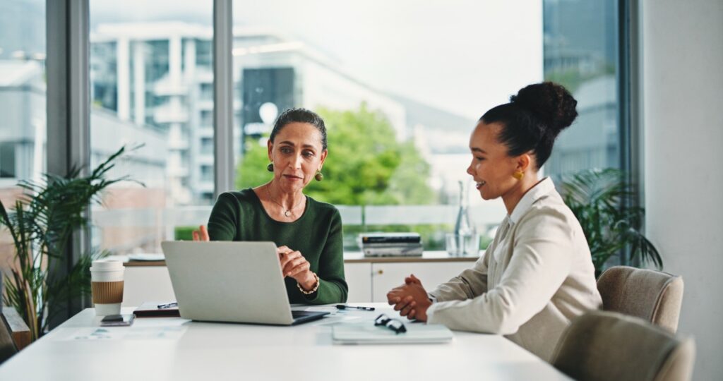 Two women sit at a table in a bright modern office, discussing work beside an open laptop. One gestures towards the laptop while the other listens. Notebooks, a coffee cup and glasses sit on the table, with large windows and plants in the background.