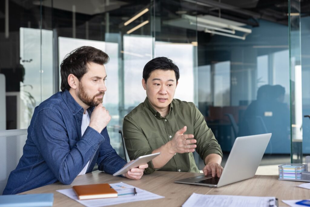 Two professionals sit at a table in a modern office, discussing information displayed on a laptop and tablet. Papers and notebooks are spread across the table as they work together.