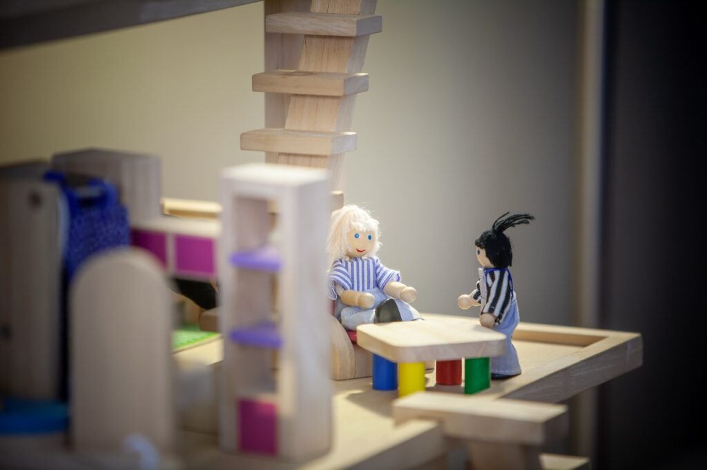 Wooden figures and building blocks arranged in a play therapy setting, with two figures seated opposite each other at a small table.