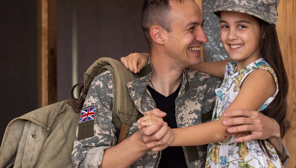 A smiling man in military uniform holds his young daughter, who has her arm around his shoulders. A Union Jack patch is visible on his uniform.