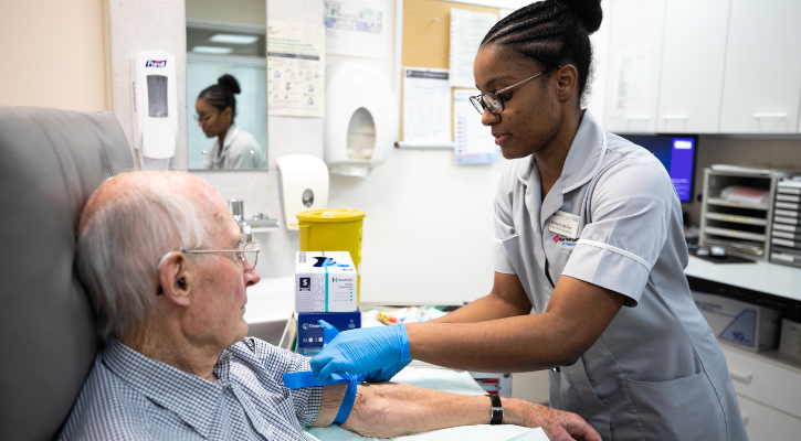 A healthcare professional wearing an NHS uniform and blue gloves takes blood from an older man seated in a clinical room at the Amyloidosis Centre at Royal Free London NHS Foundation Trust