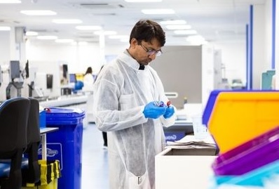 A laboratory worker wearing a white disposable gown, blue gloves and safety glasses stands at a bench, carefully examining a small sample or vial. The modern laboratory is bright and spacious, with equipment and workstations stretching into the background. Colour-coded clinical waste bins are visible nearby, while blurred laboratory containers in the foreground frame the scene, highlighting the focus on careful scientific work.