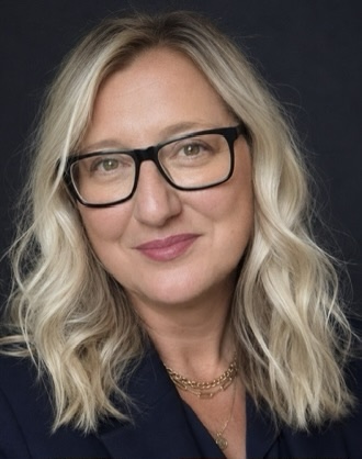 Head-and-shoulders portrait of Jo Bowers, with shoulder-length wavy blonde hair and black-framed glasses, wearing a dark navy top and layered gold necklaces against a dark background.