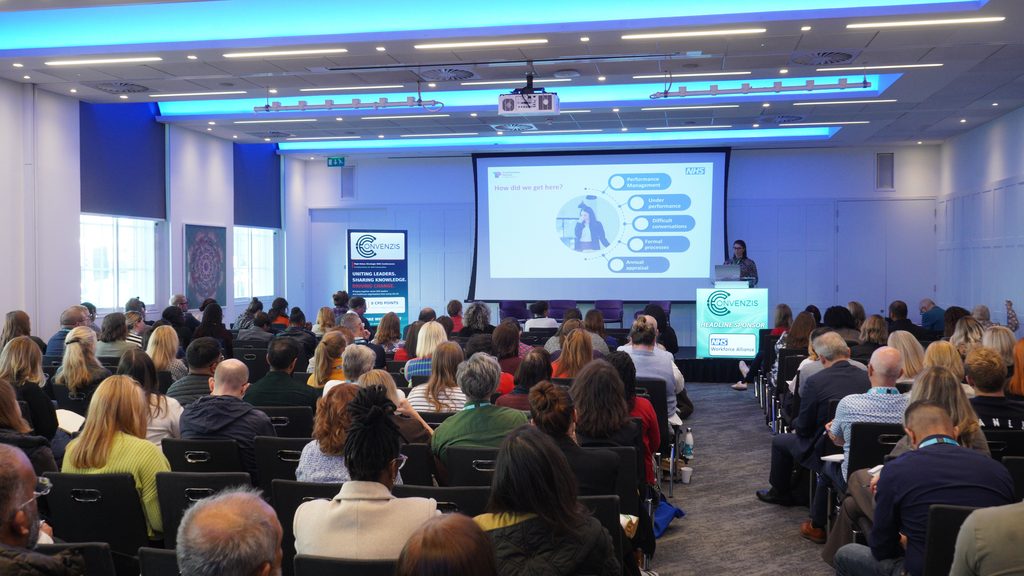 Conference room with lots of attendees sat down. Lisa presenting in front of large slide.