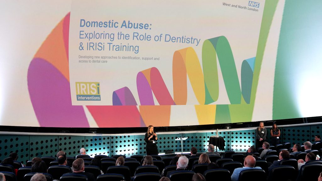 A speaker presents to an audience in a large auditorium beneath a projected slide titled “Domestic Abuse: Exploring the Role of Dentistry & IRISi Training.” The slide, branded with NHS West and North London and IRISi Interventions logos, features a colourful ribbon graphic and text about developing new approaches to identification, support and access to dental care. Attendees are seated in rows facing the stage.